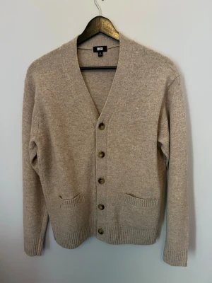 Beige kofta från Uniqlo - Mysig beige kofta från Uniqlo med v-ringning och fem knappar framtill. Koftan har två fickor fram och ribbade muddar vid ärmslut och nederkant. Perfekt att slänga på sig över en t-shirt eller skjorta. Storlek xs men sitter som s