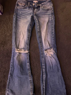 Blå bootcut jeans - Ett par blå bootcut jeans med håll på knäna, och med fickor med ett rött broderi på. 