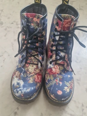 Blommiga boots från Dr. Martens - Säljer ett par unika Dr. Martens boots med blå bas och färgglatt blommönster i rosa, rött och grönt. Klassisk hög modell med svarta snören och gul söm runt sulan. Tillverkade i tyg med Dr. Martens ikoniska dragflik bak. Perfekta för dig som vill sticka ut! Storlek 38/39. Knappt använda!