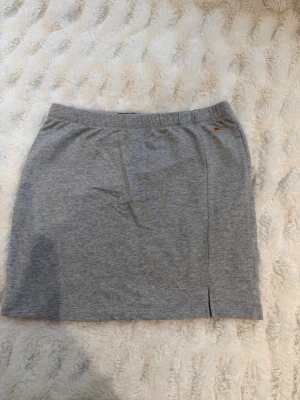 Grå kortkjol från Gant - Snygg grå kortkjol från Gant i mjuk bomull med elastisk midja och liten broderad logga framtill. Kjolen har en enkel design med en liten slits nedtill för extra rörelsefrihet. Perfekt för en avslappnad och stilren look.