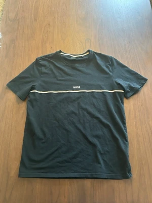 Svart t-shirt från BOSS med vit detalj - Svart t-shirt från BOSS med en vit linje och BOSS-logga över bröstet. Klassisk passform med rund hals och korta ärmar. Tillverkad i mjuk bomull som känns skön mot huden. Perfekt för en stilren och enkel look.