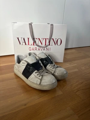 Valentino Garavani vita sneakers - Snygga vita sneakers från Valentino Garavani med en bred svart läderdetalj över sidan. Klassisk låg modell med rund tå och vita snören. (Skorna tvättas självklart innan de skickas). Priset är inte hugget i sten.