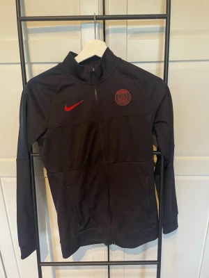 Svart PSG träningsjacka från Nike - Svart träningströja från Nike med PSG-logga och röd Nike Swoosh på bröstet. Jackan har hel dragkedja, ståkrage och diskret mönster på bröstpartiet. Perfekt för fotbollsträning eller som sportig vardagsstil. Storlek XS. Den är äkta och är köpt i Paris. Tröjan har några defekter bland annat hål i fickan. 