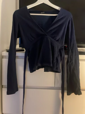 Mörkblå omlottblus med långa ärmar från Gina storlek xs - Snygg mörkblå omlottblus från Gina Tricot med långa ärmar och knytband i midjan. Blusen har en djup v-ringning och är tillverkad i ett mjukt, stretchigt tyg som sitter bekvämt. .