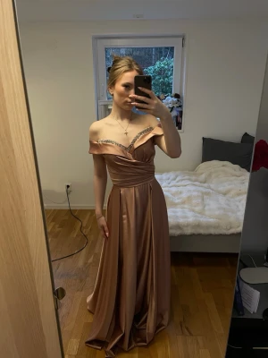 Champagnefärgad långklänning med paljettdetalj - En jättefin och elegant festklänning i satin för dam i storlek 36-38. Klänningen har en rosa guld färg och är axelbandlös, men det går att lägga band om man vill ha. Köpt i MK Mode i Södertälje. Klänningen har en omlottdetalj över bysten och dekor längs urringningen. Modellen är lång med ett bra lager och har ett till tunt lager under markers midja. Perfekt för bal eller festliga tillfällen.