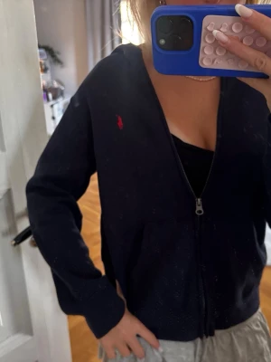 Mörkblå hoodie från Ralph Lauren - Snygg mörkblå hoodie med dragkedja från Ralph Lauren. Klassisk design med röd broderad logga på bröstet och huva. Perfekt för en chill och stilren look. Mjukt material och bekväm passform.
