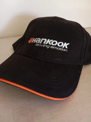 Svart Hankook keps med brodyr - Svart keps från Hankook med broderad logga och texten 'driving emotion' framtill. Kepsen har böjd skärm med orange kant och klassisk sexpanelsdesign. Perfekt accessoar för bilentusiaster eller motorsportfans.