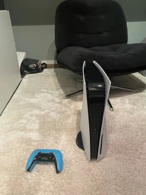 PlayStation 5 med blå DualSense-kontroll - PlayStation 5 spelkonsol från Sony med futuristisk vit och svart design. Medföljer en blå DualSense trådlös handkontroll. Konsolen har en stilren, stående form och är tillverkad i plast med matta och blanka ytor. Perfekt för gaming på nästa nivå.