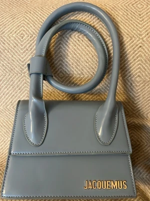 Blå miniväska från Jacquemus - Säljer en stilren blå miniväska från Jacquemus i glansigt eco  läder. Väskan har ett elegant, fyrkantigt format med lång axelrem och guldfärgad Jacquemus-logga framtill. Perfekt för dig som gillar trendiga accessoarer och vill sticka ut med en ikonisk design. 