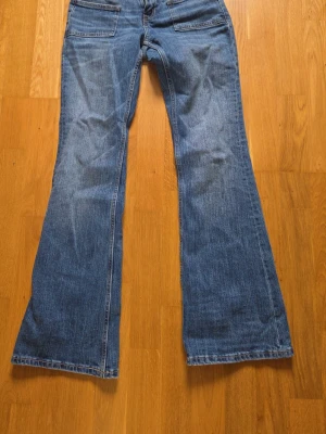 Blå bootcut jeans med fickor fram - Säljer ett par blå bootcut jeans med två stora fickor framtill och klassisk knappgylf. Jeansen har en snygg tvätt och markerade sömmar. Perfekta för dig som gillar retrostil och vill ha ett par jeans med lite vidare ben nedtill.