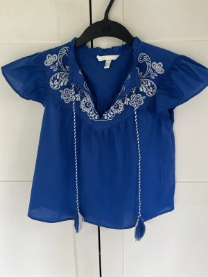 H&M - Supersöt blå blus från H&M med vita broderier och knytband med tofsar. Blusen har korta ärmar och ett luftigt, lätt material. Snygg v-ringning och fina detaljer vid halsen.