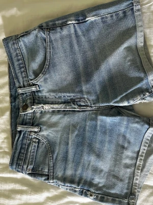Ljusblå denimshorts från Levi's - Säljer ett par klassiska ljusblå denimshorts från Levi's med fem fickor och råa kanter. Shortsen har normal passform och är perfekta för sommaren. De har en knappgylf och bälteshällor för enkel styling.