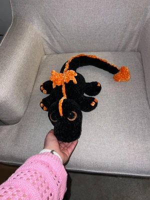 Svart drake med orangea detaljer - Unik handgjord salamander i mjukt, svart garn med orange detaljer och stora glittriga ögon. Draken har en lång svans, fyra ben och ett dekorativ hjärta på svansen. Perfekt som dekoration eller mysig maskot.🧡🧡