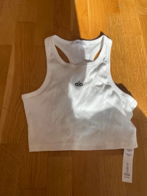 Vit ribbad linnetopp från Alo - Snygg vit ribbad linnetopp från Alo med brottarrygg och diskret logga framtill. Toppen är croppad och har en sportig känsla, perfekt till jeans eller träningstights. Köpt från en annan tjej, så jag är inte helt säker på om den är äkta.