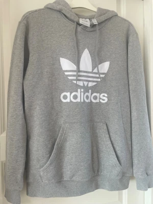 Grå hoodie från Adidas Originals - Säljer en grå hoodie från Adidas Originals med stor vit logga och text framtill. Tröjan har huva med dragsko och en stor magficka. Tillverkad i mjukt sweatshirtmaterial, perfekt för chill eller träning.