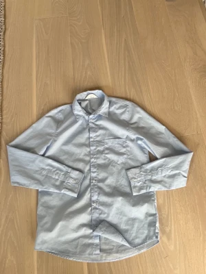 Ljusblå skjorta HM - Snygg ljusblå skjorta med klassisk krage och bröstficka. Skjortan har långa ärmar och knäppning framtill. Perfekt för en stilren look och enkel att matcha med olika outfits. Passar 170cm