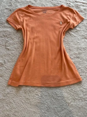 Korall t-shirt från Polo Ralph Lauren - Snygg korall färgad t-shirt från Polo Ralph Lauren med klassisk passform och liten broderad logga på bröstet. Den är i storlek 150 12-14 år men passar som en xs. Den ser orange ut på bilden men är i verkligheten mer rosa/korall färgad.💕