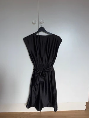 Svart satinklänning med knyt i midjan - Svart klänning från H&M i glansigt satinmaterial. Modellen har kort ärm, markerad midja med knytband och rundad halsringning. 