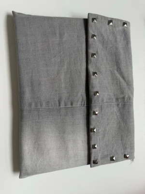 Grå datorfodral med nitar - Stilren grå datorfodral i tyg med silverfärgade nitar längs kanten. Väskan har en enkel, rektangulär form och stängs med ett lock. Perfekt accessoar för att addera edge till din outfit.