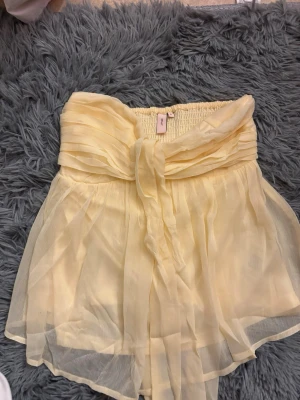 Gul chiffong bandeau topp med rosett - Supersöt ljusgul bandeau topp i chiffong med stor rosett framtill. Toppen är axelbandslös och har ett flowy, luftigt tyg som ger en romantisk känsla. Perfekt för varma dagar och somriga outfits. Aldrig använd endast testad!!