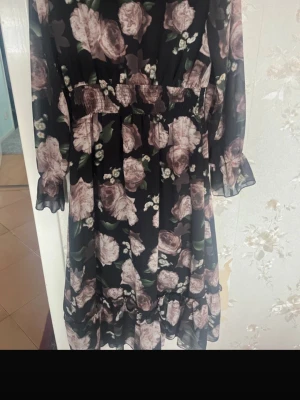 Svart blommig långklänning - Svart långklänning med stora rosa och ljusa blommor. Klänningen har långärmade, transparenta ärmar och smock i midjan för en snygg passform. Volangdetaljer längst ner och vid ärmsluten ger extra stil. Perfekt för dig som gillar romantiska mönster.