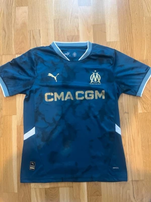 Marseille 24/25 away/bortatröja - Snygg Marseille matchtröja från Puma i mörkblått med guldiga detaljer och logga. Tröjan har korta ärmar, vit kant vid ärmslut och krage samt tryckt sponsor på bröstet. Tillverkad i lätt och ventilerande material, perfekt för fotbollsträning eller match. Tröjan är Authentic. Tröjan är i storlek S