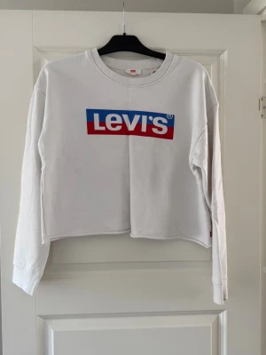 Vit Levi's hoodie med logga - Säljer en vit hoodie från Levi's med stor färgglad logga i blått, rött och vitt på bröstet. Tröjan har långa ärmar, rund halsringning och en avslappnad passform. Perfekt för en casual och trendig stil.