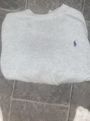 Ljusgrå stickad tröja från Ralph Lauren - Snygg ljusgrå stickad tröja från Ralph Lauren med klassisk rund halsringning och den ikoniska blå loggan broderad på bröstet. Perfekt för dig som gillar stilrena och tidlösa plagg. Tröjan är ribbstickad vid halsen och har en mjuk känsla.