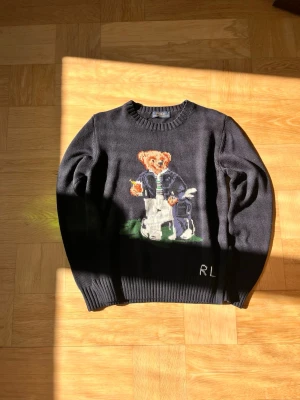 Mörkblå stickad tröja Polo Bear - Mörkblå stickad tröja från Polo Ralph Lauren med ikoniskt Polo Bear-motiv framtill. Tröjan har rund halsringning, ribbstickade muddar och RL-broderi vid nederkanten. Perfekt för dig som gillar klassisk och lekfull stil.