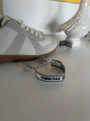 Christian Dior J'Adior armband svartvit - Snyggt armband från Christian Dior i svart och vitt med texten 'J'Adior'. Armbandet är vävt i tyg och har justerbara tofsar för en cool och personlig look. Perfekt accessoar för att lyfta din outfit med en lyxig touch.