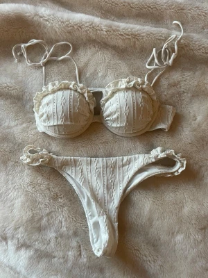 Vit bikini med volang från Nelly - Supersöt vit bikini från Nelly med volangdetaljer och knytband. Bikinin har ett subtilt mönster i tyget och push up-topp med vadderade kupor. Perfekt för strandhäng och sommarens alla bad.