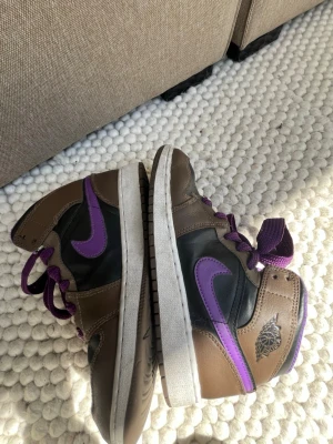 Nike Air Jordan 1 High brun/lila - Säljer ett par Nike Air Jordan 1 High sneakers i brunt och svart med lila detaljer och lila snören. Skorna har klassisk hög siluett, vit sula och ikonisk Air Jordan-logga på sidan. Perfekta för dig som vill sticka ut med färg och stil.