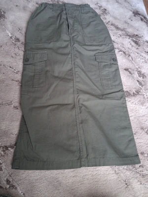 Grön cargokjol med fickor - Stilren lång cargokjol i militärgrönt med stora sidofickor och knappdetaljer. Kjolen har rak passform och är tillverkad i ett slitstarkt bomullsmaterial. Perfekt för en avslappnad och trendig look. Passar även dig som bär xs/s 😃