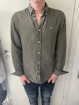 Olivgrön linneskjorta från Morris - Stilren olivgrön skjorta från Morris i 100% linne. Skjortan har klassisk krage, vita knappar och en liten broderad logga på bröstet. Perfekt för dig som gillar en avslappnad men snygg look. Skjortan har mindre fläck under armålan men det syns knappt och är inget man tänker på. Modellen på bilden är 185 cm och väger 75 kg.