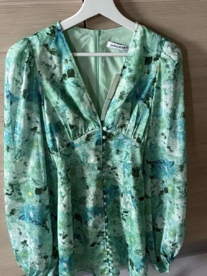 Blommig grön kort klänning från Adoore - Supersnygg kort klänning från Adoore i gröna och blå toner med blommigt mönster. Klänningen har v-ringning, långa puffärmar och knappar framtill. Tillverkad i ett lätt, glansigt material som känns lyxigt och flowy. Klänningen är oanvänd och prislappen sitter kvar🤍 