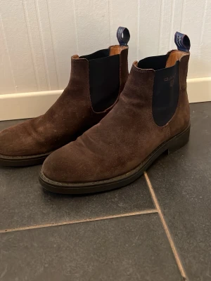 Bruna Chelsea boots från Gant - Säljer ett par snygga bruna Chelsea boots från Gant i mocka. Skorna har elastiska sidopaneler i mörkblått med Gant-logga och dragflikar bak. Klassisk siluett med robust sula och låg klack. Perfekta för dig som gillar stilrena och tidlösa boots.