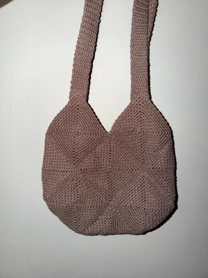 Gråbrun virkad påse - Säljer en gråbrun påse som är virkad i ett snyggt geometriskt mönster. Den har breda axelband och en mjuk, rundad form. Påsen mäter 25x25 och snörena är cirka 73cm långa.