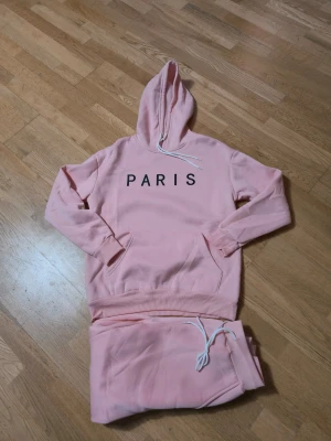 Rosa mjukisbyxor med hoodie, PARIS - Säljer ett set med rosa mjukisbyxor och matchande hoodie med texten PARIS på bröstet. Byxorna har dragsko i midjan och muddar vid bensluten. Hoodien har känguruficka och vit dragsko i huvan. Perfekt för chill dagar eller när du vill vara extra comfy.
