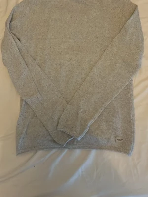 Ljusgrå stickad tröja Jack & Jones  - Ljusgrå stickad tröja från Jack & Jones med rund halsringning och långa ärmar. Tröjan har en enkel, rak passform och är tillverkad i mjukt material. Liten diskret logolapp nertill på framsidan. Perfekt basplagg för lager på lager.