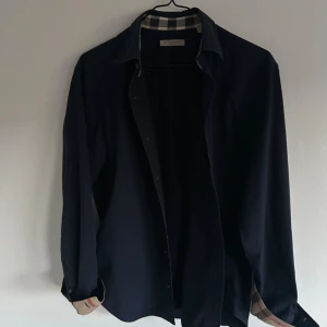 Mörkblå skjorta med rutiga detaljer - Stilren mörkblå skjorta från Burberry med klassiska beige-rutiga detaljer på insidan av kragen och ärmsluten. Skjortan har lång ärm, knappar framtill och en normal passform. Perfekt för dig som gillar tidlös design med en twist.