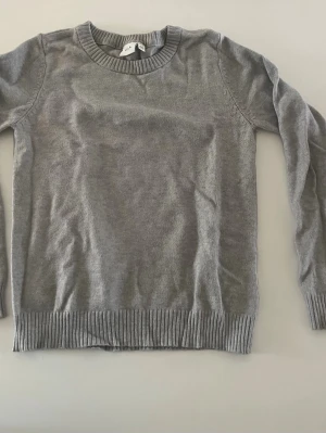 Grå stickad tröja  - En enkel och stilren grå stickad tröja med rund halsringning och ribbade muddar vid ärmslut och nederkant. Perfekt basplagg att matcha med jeans eller kjol. Materialet känns mjukt och lätt, vilket gör den skön att bära hela dagen.