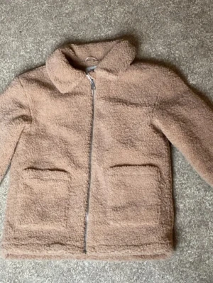 Beige teddyjacka från KappAhl - Mysig beige teddyjacka från KappAhl med dragkedja framtill och två stora fickor. Jackan har klassisk krage och är gjord i fluffigt teddy-material som håller dig varm och trendig. Perfekt för dig som gillar en avslappnad och cool stil.