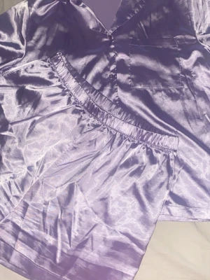 Ljuslila satinpyjamas set - Snyggt pyjamasset i ljuslila satin med kortärmad skjorta och matchande shorts. Skjortan har knappar framtill och en bröstficka, shortsen har elastisk midja för extra komfort. Perfekt för mysiga kvällar hemma.