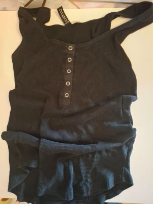 Svart ribbad linnetopp med vingar - Säljer en svart ribbad linnetopp från Collusion med tryckta änglavingar i metallicfärger på ryggen. Toppen har knappdetaljer framtill och är ärmlös med breda axelband. Perfekt för dig som gillar edgy och unik stil.