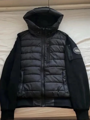 Svart pufferjacka Supply & Demand - Snygg svart pufferjacka från Supply & Demand med huva och quiltad design. Jackan har dragkedja framtill, fickor med dragkedja och en patch på ärmen. Perfekt för kalla dagar och ger en modern streetwear-look.