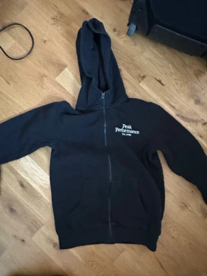 Svart zip-hoodie Peak Performance - Svart hoodie med dragkedja från Peak Performance. Klassisk modell med huva, fickor framtill och vit logga på bröstet. Perfekt för en avslappnad och sportig stil. Mjuk bomullskänsla och enkel att matcha till det mesta.
