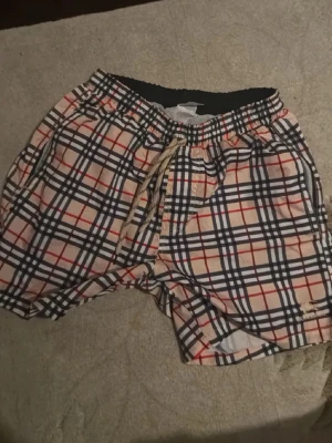 Rutiga badbyxor från Burberry - Snygga badbyxor från Burberry med klassiskt rutmönster i beige, svart, vitt och rött. De har resår i midjan med snörning och sidofickor. Materialet är lätt och snabbtorkande, perfekt för stranden eller poolen. Burberry-logga broderad på benet.
