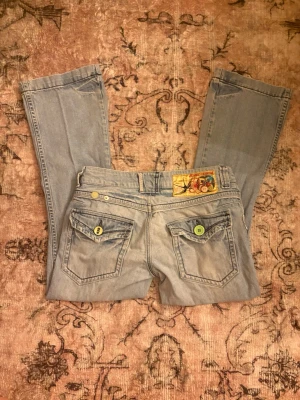 Ljusblå bootcut jeans med färgglada detaljer - Unika ljusblå jeans med bootcut passform och två bakfickor med färgglada knappar. Snygg broderad patch ovanför fickan bak ger extra karaktär. Perfekta för dig som vill sticka ut lite extra. Klassisk denimkänsla och cool vintage vibe.