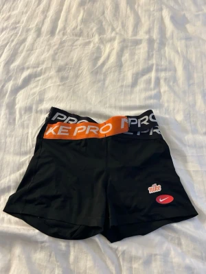 Svarta Nike Pro träningsshorts - Säljer ett par svarta Nike Pro träningsshorts med elastisk midja i svart och orange med vit text. Shortsen har en tight passform och är tillverkade i ett stretchigt syntetmaterial, perfekta för träning. Nike-logga i rött och vitt framtill.