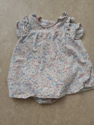 Babyklänning - Supersöt babyklänning i mjuk bomull med blommigt mönster i pastellfärger som rosa, blått och vitt. Klänningen har volangärmar och inbyggd body undertill för extra bekvämlighet. Perfekt för varma dagar och har en klassisk, gullig look.